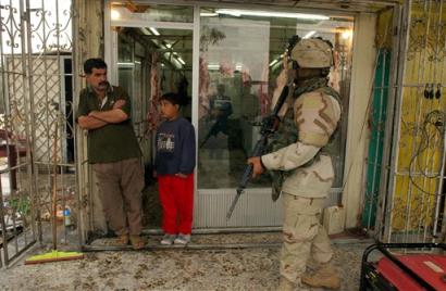 An Iraqi and his son stand outside their shop in Baghdad's Kadesia district as a US soldier walks past Wednesday Dec 8, 2004. US troops searched houses for weapons in the neighborhood. [AP]