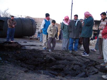 People look at a crater left when a natural gas tanker exploded in Mahoumdiya, some 25 kilometers (15 miles) south of Baghdad, Iraq, Thursday Dec. 23 2004, killing four people and injuring about 50, according to hospital director Dr. Dawoud al-Taei. The tanker, seen in background left, was en route to Baghdad when, according to witnesses, it was hit by a rocket and exploded. [AP]