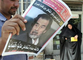 A woman stands next to a man reading Iraqi newspapers bearing a photo of Saddam Hussein on the front page, in Baghdad, Iraq, Monday, Sept. 5, 2005 with the headline referring to the impending trial.