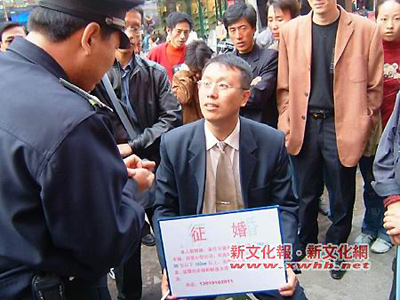 A young man sits on the street holds an advertisement for himself waiting for his own "Mrs Right" with curious people surrounding him.