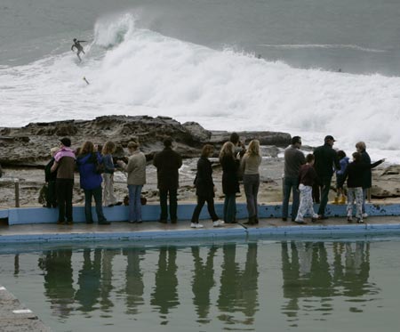 Onlookers are reflected in a saltwater swimming pool as they watch a surfer ride a wave at Dee Why Point in Sydney June 10, 2007. The death toll from a storm and torrential rain hitting Australia's east coast stands at seven with two still missing, said police as forecasters say conditions will begin to ease.