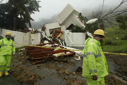 Los Angeles utility workers Doug Quintal, left, and Eric Lawrence pass a two-story house that collapsed after a mudslide Monday Jan 10, 2005, in the Coldwater Canyon section of Los Angeles. Authorities said a father and his two children were pulled from the rubble of the home. The father had minor injuries. Nine deaths have been linked to a series of storms pounding Southern California that have unleashed flash floods and mudslides, forced evacuations and closed roads and schools. [AP]