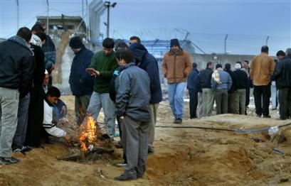 Before sunrise, Palestinian workers warm themselves by the fire waiting for the opening of the Israeli-run industrial complex, visible in background, on Israel's border with northern Gaza Strip, at Erez, Sunday, Feb. 13, 2005. After months of closure, Israel has reopened the complex due to the current official ceasefire with the Palestinian Authority. [AP]