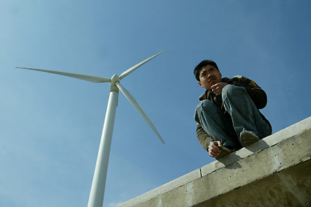 Chinese people stand behind the wall of a windmills site, used as an energy source, at Feng Xian in Shanghai, China February 24, 2006. China, faced with a growing shortage of energy as the economy gallops forward, is looking to add energy conservation as a criterion of officials' performance reviews, a planning official said on Thursday.