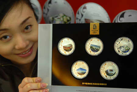 A saleswoman shows a set of commemorative badges featuring five of the venues for the Beijing Olympic Games: (from top left to bottom right ) the National Stadium, or 'Bird's Nest', Qingdao Olympic Sailing Center, Hong Kong Olympic Equestrian Venue, National Aquatics Center, or 'Water Cube', and Shenyang Olympic Stadium , in Beijing, March 16, 2008. [Xinhua]