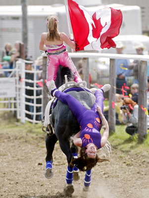 Trick rider Shelby Cummings stands carrying the Canadian flag while another trick rider Meghan Gulliver hangs from the back of a galloping horse at the 53rd annual Water Valley Rodeo, near Water Valley, Alberta, June 4, 2006. [Reuters]